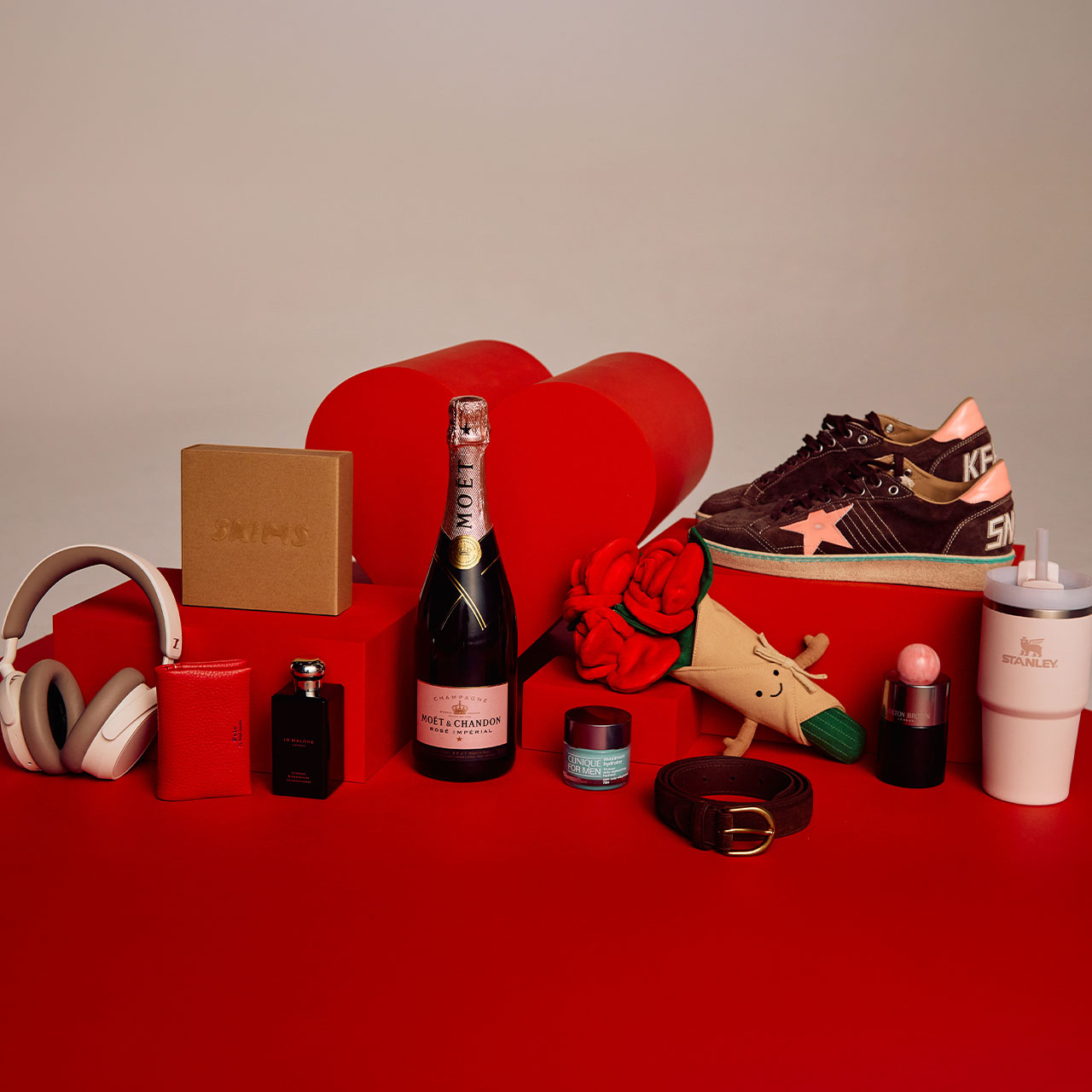 A collection of gifts including headphones, a bottle of champagne, sneakers, cosmetics, a plush rose, and a branded tumbler on a red surface.